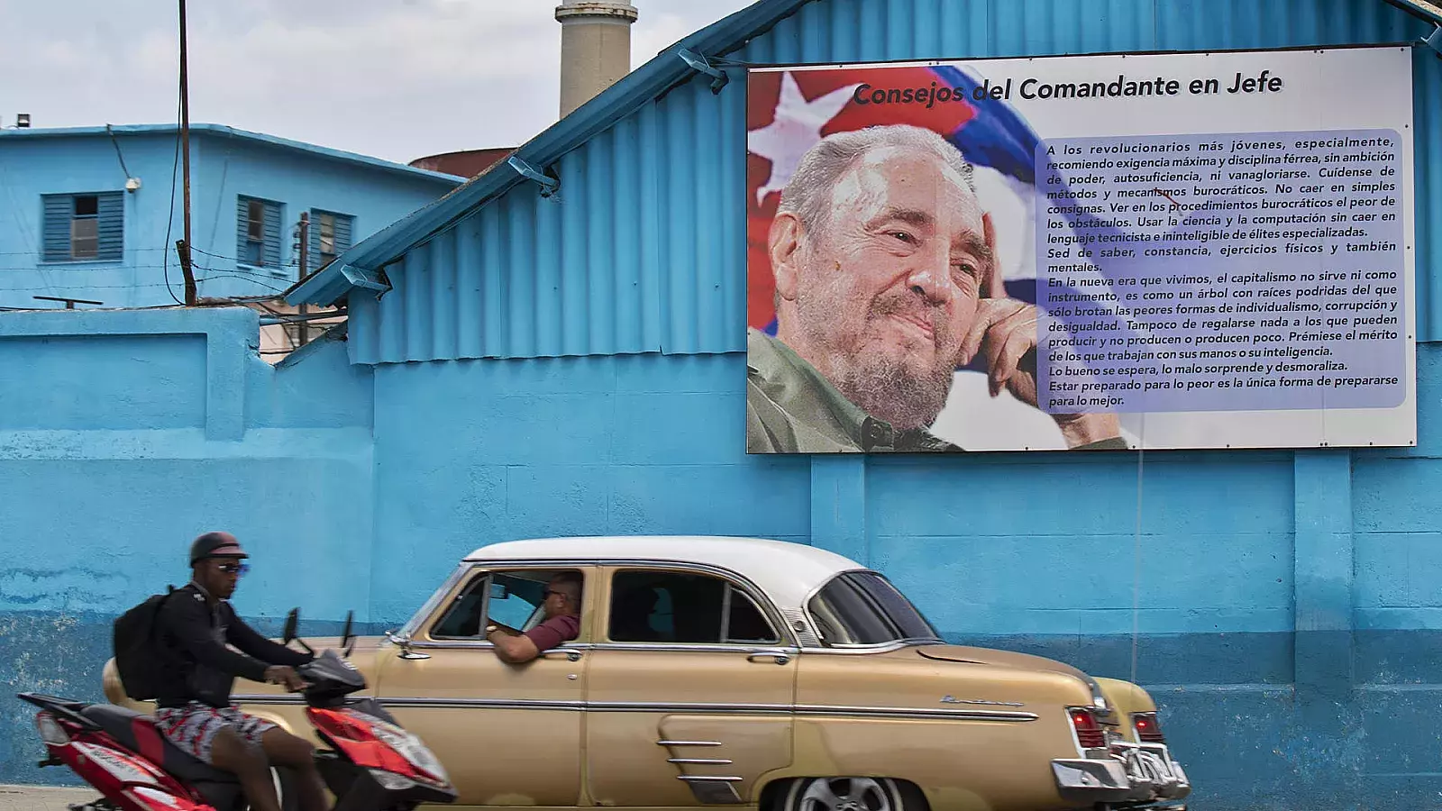 Un auto y una motocicleta pasan frente a una valla con un retrato de Fidel Castro en La Habana
